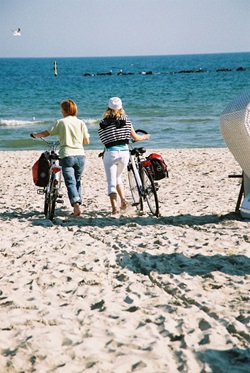 2 Radlerinnen schieben ihre Räder über einen Sandstrand auf die Ostsee zu.