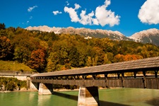 Eine überdachte Holzbrücke überquert einen grünlich schimmernden Fluss; im Hintergrund erstrecken sich ein herbstlich gefärbter Wald und ein eindrucksvolles Alpenpanorama.