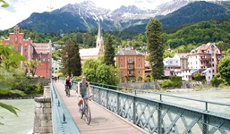 Zwei Radfahrerinnen auf der Innsteg-Brücke in Innsbruck - im Hintergrund ein traumhaftes Bergpanorama und die St. Nikolaus-Kirche.