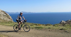 Ein Mountainbiker fährt einen Weg entlang der Südlichen Ägäis - im Hintergrund ist das tiefblaue Meer zu sehen