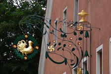 Ein geschmiedetes Schild mit einem Hahn, der vergoldet ist, an einer rölichen Hauswand