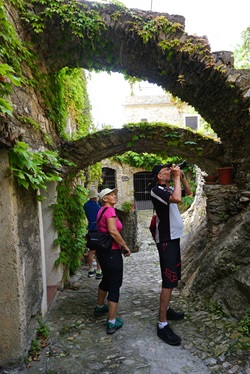 Zwei Radtouristen bummeln durch die romantischen, teilweise mit Efeu bewachsenen Bogengänge eines ligurischen Dorfes und machen Fotos.