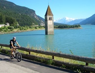 Ein Radfahrer fährt auf dem Etschtal-Radweg am Reschensee und dem versunkenen Kirchturm von Graun vorbei.
