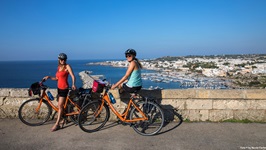Zwei Radlerinnen posieren an einer Küstenstraße im Salento für ein Foto.