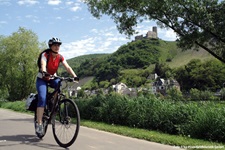 Eine Radlerin fährt auf dem Mosel-Radweg entlang, im Hintergrund sind eine Burgruine und ein Dorf zu sehen
