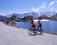 Zwei Radlerinnen fahren auf der Passhöhe des St. Gotthard am Seeufer entlang auf das Gotthard-Hospiz und das St-Gotthard-Museum zu. Im Hintergrund erheben sich schneebedeckte Alpengipfel.