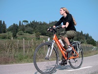 Eine Radfahrerin fährt auf einer Straße an einem mit Zypressen bewachsenen Hügel in der Toskana entlang.