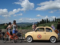 Zwei Radfahrerinnen und weitere Touristen machen eine Pause am Straßenrand und genießen den Ausblick auf die von Zypressen geprägte Hügellandschaft der Toskana.