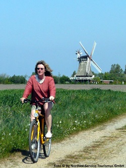 Eine Fahrradfahrerin fährt auf einem Schotterweg in Nordfriesland entlang, im Hintergrund ist eine Windmühle zu sehen