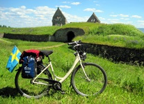 Ein Fahrrad mit Gepäcktasche und einer schwedischen Fahne steht inmitten einer grünen Wiese vor einer Burgruine mit markanten Treppengiebeln.