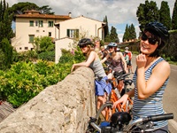 Mehrere Radlerinnen machen in einem toskanischen Dorf Pause und genießen, an eine Steinmauer gelehnt, die Aussicht.