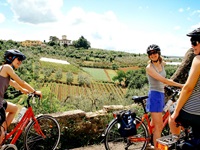 Drei Radlerinnen in der Toskana machen bei einem Steinmäuerchen Pause und genießen den Blick auf die umgebenden Weinberge.