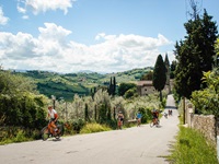RadfahrerInnen auf einem asphaltierten, von Zypressen und Steinmäuerchen gesäumten Radweg in der Toskana. Im Hintergrund ist der Kirchturm eines Dorfes zu erkennen.