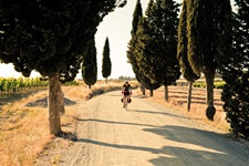 Eine Radlerin in der Toskana folgt einem geschotterte Radweg durch eine Zypressenallee.