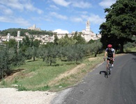 Ein Radfahrer auf einer Straße bei Assisi, im Hintergrund die Silhouette der Stadt.