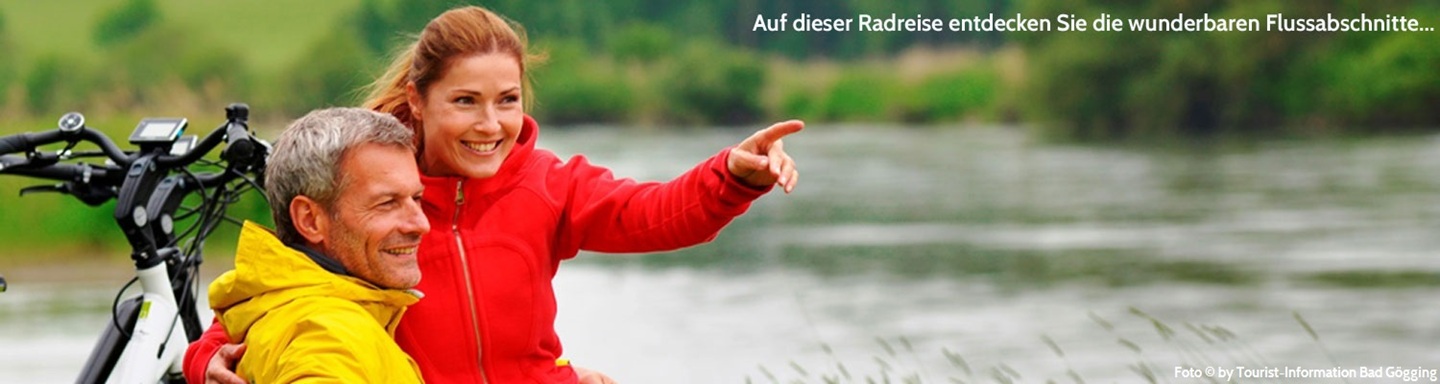 Ein Radlerpaar, sie mit roter und er mit gelber Jacke neben einem Fluss in Bad Gögging, sie zeigt auf etwas in die Ferne