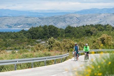 Zwei Radfahrer radeln auf einer asphaltierten Straße nebeneinander über die Insel Hvar. Im Bildhintergrund sind das Meer und eine Bergkette zu erkennen.