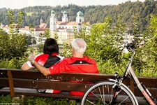 Ein Pärchen sitzt oberhalb von Passau auf einer Bank und genießt den Blick auf den Passauer Dom.