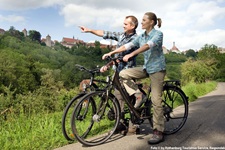 Zwei Radfahrer auf einem geteerten Weg halten am Flussufer bei Rothenburg - ein Mann zeigt mit seinem Finger auf etwas