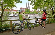 Zwei Radlerinnen stehen an der Donau und betrachten den Regensburger Dom auf der gegenüberliegende Flussseite.