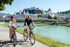 Zwei Radfahrer radeln an der Salzach entlang, im Hintergrund die Altstadt von Salzburg mit dem Dom und der Festung Salzburg.