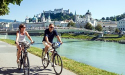 Zwei Radfahrer radeln an der Salzach entlang, im Hintergrund die Altstadt von Salzburg mit dem Dom und der Festung Salzburg.