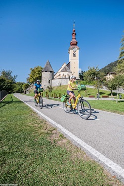 Zwei Radler auf dem Alpe-Adria-Radweg passieren die Kirche St. Peter und Paul in Tarvis.