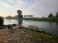 Ein Fahrrad steht an einem befestigten Ufer eines Flusses, an dem ein kleiner Steg zu einem angelegten Boot führt
