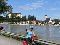 Eine Radfahrerin fährt auf dem Donauradweg, winkt in die Kamera, neben sich die Donau und dahinter eine Stadt.