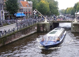 Blick über den Kanal mit einem Boot in Amsterdam zur Promenade, auf der zahlreiche Fahrräder abgestellt sind#