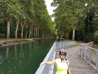 Eine Radlerin posiert auf einer Brücke im Elsass, welche auf einen Weg durch einen mit am Ufer gepflanzten Birken führt
