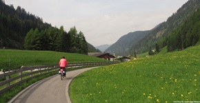 Eine Radllerin fährt auf einem von Wiesen gesäumten Radweg auf ein kleines Gehöft zu.