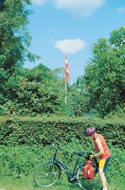 Landschaft in Dänemark mit der dänischen Flagge im Hintergrund - eine Radlerin holt ihre Kamera aus ihrer Gepäcktasche um dieses zu fotografieren
