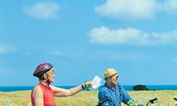 Ein Radlerpaar steht auf einem Radweg neben Feldern, die Frau zeigt in eine Richtung, während sie die Routenbeschreibung in der Hand hält