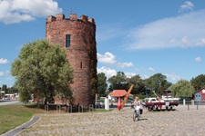 Eine Radfahrerin fährt auf Kopfsteinpflaster an einem zinnenbewehrten Backsteinturm vorbei.