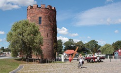 Eine Radfahrerin fährt auf Kopfsteinpflaster an einem zinnenbewehrten Backsteinturm vorbei.