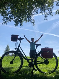 Ein Fahrrad wurde unter einem Baum auf einer Wiese abgestellt, dahinter steht die Radfahrerin und posiert mit ausgestreckten Armen für die Kamera