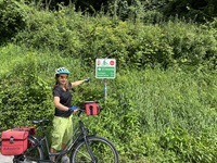 Eine Frau steht neben ihrem Fahrrad und zeigt auf einen Wegweiser des Donauradwegs, der in Richtung Schlögen und Niederranna führt