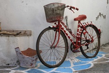 Impression der südlichen Griechischen Ägäis: Ein altes Fahrrad in rot-weißer Farbe und einem Fahrradkorb ist an einer Häuserwand angekettet - davor steht eine zerbrochene, weiß bemalte Teracotta-Vase. Der Boden ist in blau und weißer Farbe angemalt