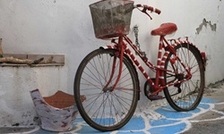 Impression der südlichen Griechischen Ägäis: Ein altes Fahrrad in rot-weißer Farbe und einem Fahrradkorb ist an einer Häuserwand angekettet - davor steht eine zerbrochene, weiß bemalte Teracotta-Vase. Der Boden ist in blau und weißer Farbe angemalt
