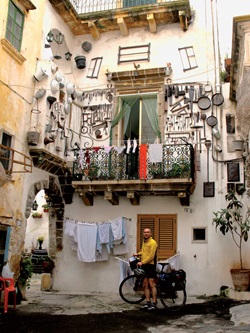 Ein Radler steht vor einem individuell mit Töpfen, Schüsseln und Eimern geschmückten Haus in Gallipoli.