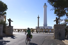 Ein Radfahrer im Salento lässt den Leuchtturm von Santa Maria di Leuca hinter sich und fährt zwischen zwei Engelstatuen hindurch auf die Basilica Santa Maria de Finibus Terrae zu.