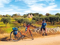 Ein Radler hat es sich für eine Trinkpause auf einem Steinmäuerchen im Salento gemütlich gemacht. Im Hintergrund stehen zahlreiche Olivenbäume und mehrere Trulli mit den typischen kegelförmigen Dächern.