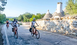 Drei Radfahrer fahren auf einem asphaltierten Radweg an einer weißen Steinmauer entlang, hinter der sich einige der für Apulien typischen Trulli erheben.