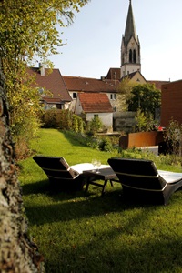Gartenbereich mit zwei schwarzen Liegen, die mit weißen Polstern belegt sind und einem kleinen schwarzen Tischchen mit zwei Weingläsern dazwischen mit Blick auf die Gebäude und den Kirchturm in Münsingen