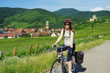 Eine Radlerin auf der Elsässischen Weinstraße bei Kaysersberg; im Bildhintergrund sind die Heilig-Kreuz-Kirche und Burg Kaysersberg zu erkennen.