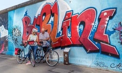 Ein Stück der ehemaligen Berliner Mauer, die mit einem Graffitti geschmückt ist und vor der zwei Fahrradfahrer stehen und in ein Prospekt schauen