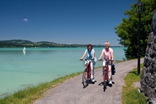 Zwei Fahrradfahrerinnen radeln einen Radweg am Forggensee entlang, auf dem ein Surfer zu sehen ist