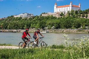 Zwei Radfahrer radeln an der Donau in Bratislava entlang, während die Burg Bratislava im Hintergrund zu sehen ist