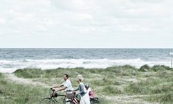 Eine kleine Familie schiebt ihre Räder durch die Dünen Dänemarks, das Kind sitzt auf dem Gepäckträger eines Fahrrads, im Hintergrund ist das Meer mit brechenden Wellen zu sehen